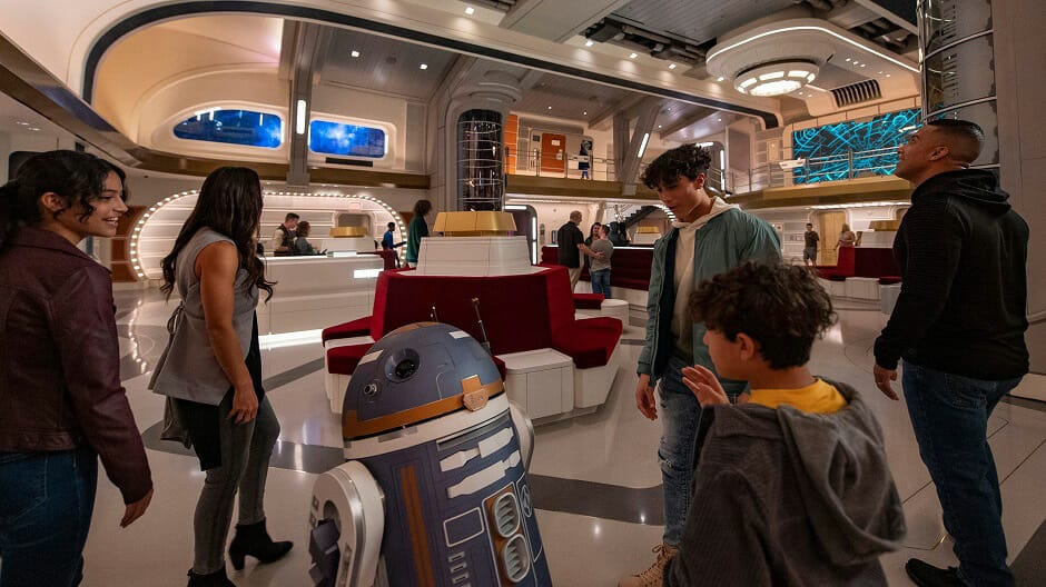 A family gazing around thefuturistic interior of the Starcruiser next to an Astromech droid SK-620